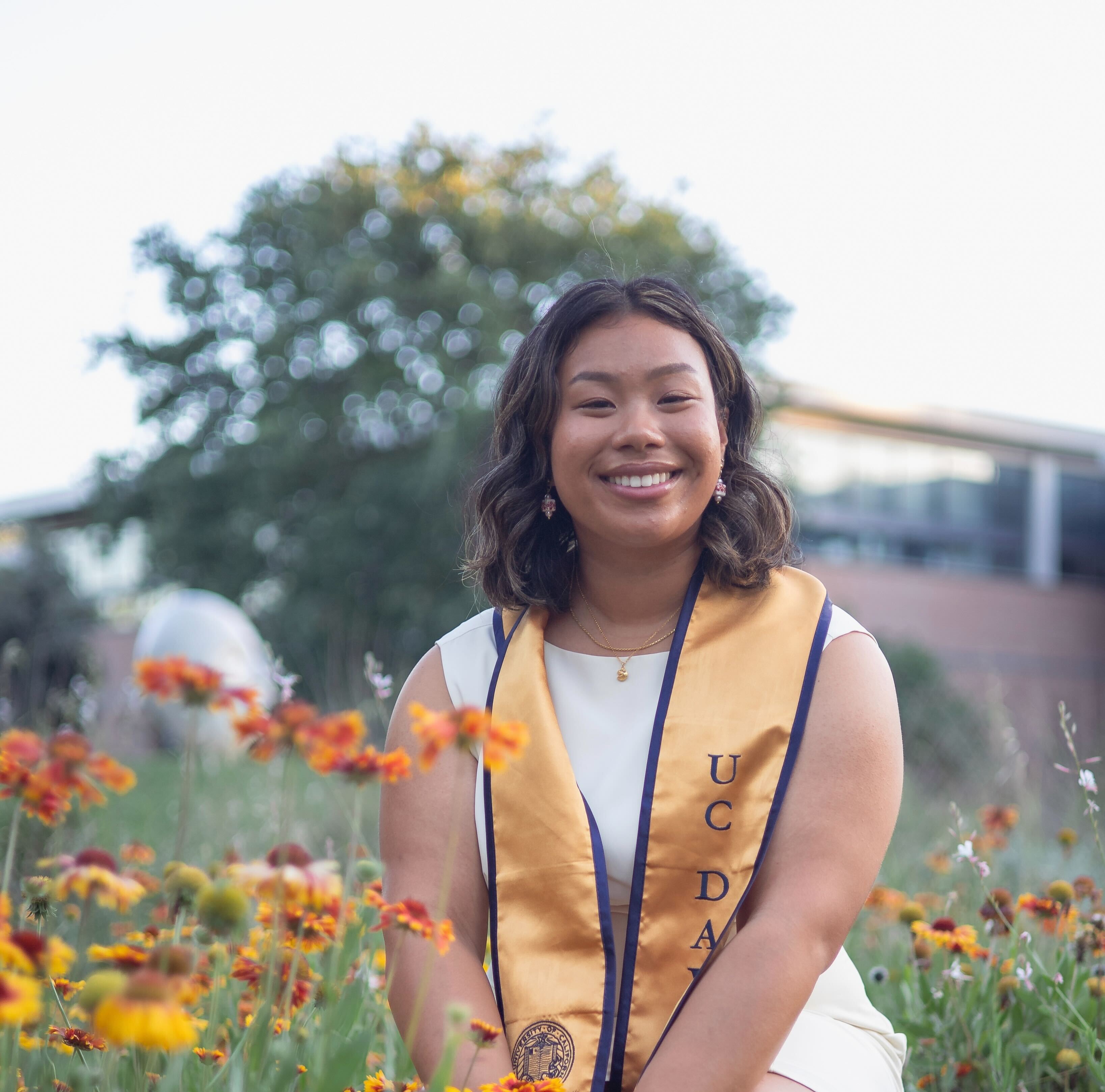 Terilyn Wong smiles, kneeling among flowers and wearing a UC Davis stole