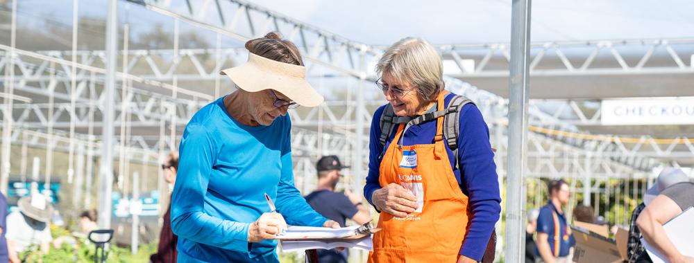 An Arboretum plant sale volunteer talking to a customer and reviewing a list on a clipboard together.