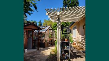 Mary Kay Williams' backyard showing a partially shaded patio area with shelving to store gardening supplies, and a small wooden shed next to a small wood and glass greenhouse in the background.