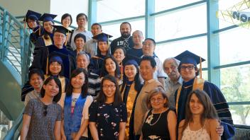A group of two dozen people, some wearing commencement caps and gowns, pose on a staircase
