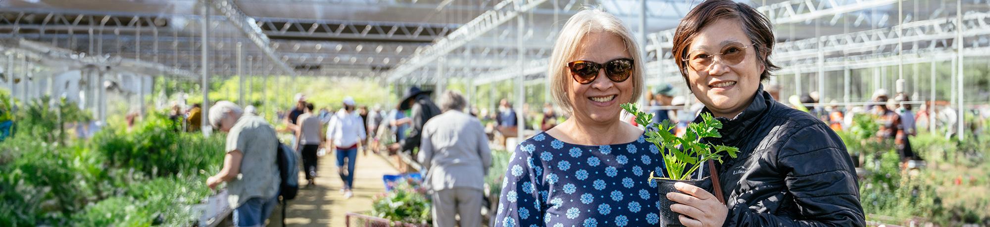 Two customers hold up plants and smile at the camera at plant sale in the UC Davis Arboretum and Public Garden Teaching Nursery.