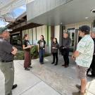 Group of six accreditation team members standing outdoors near a building, with two students, Dyanna Perez and Amelia Swanson, explaining something to the group in front of a modern, green-accented building.
